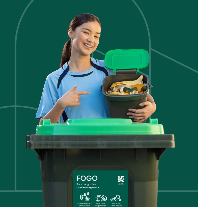 A young girl in blue shirt holding small FOGO benchtop bin with food scraps above a green lid OGO wheelie bin